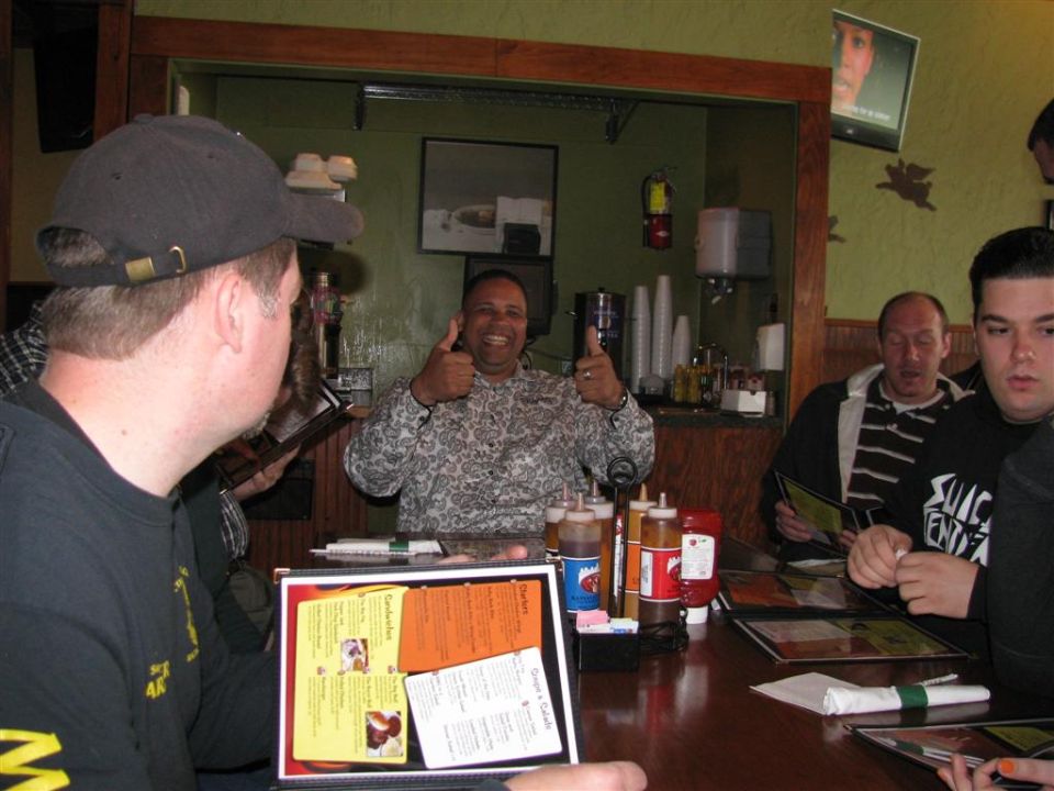 People sit at a restaurant table with menus and condiments; one man gives a double thumbs-up