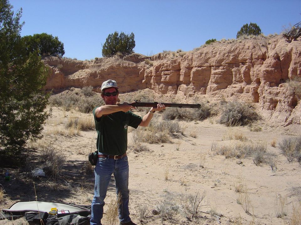 Man in jeans and t-shirt aims a rifle in a desert with rocky hills behind him. Supermotors.net logo in lower right