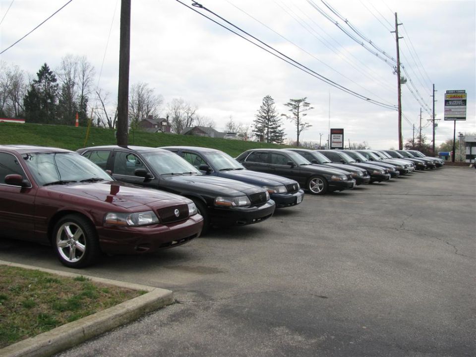 Maroon sedan in foreground, followed by a line of dark sedans parked on asphalt