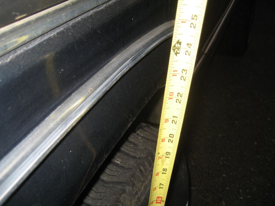 Yellow tape measure held against dark vehicle body panel near fender flare and tire, showing around 20 inches