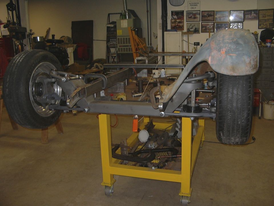 Bare car frame with front suspension and wheels on a yellow shop cart in a garage