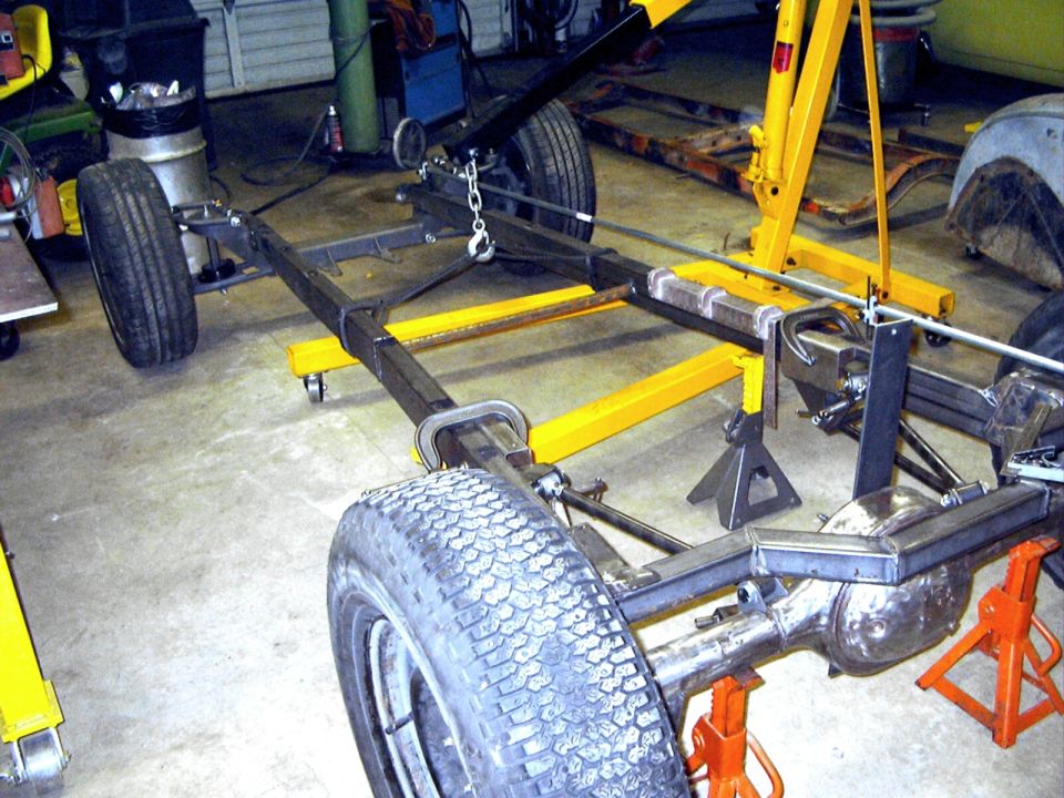 Bare vehicle chassis on jack stands and yellow jig in a shop, rear axle welded, engine hoist above
