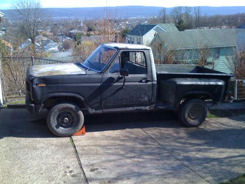 1985 Ford F-150 - A black Ford F-150 pickup truck is parked on a concrete driveway with a scenic town in the background.