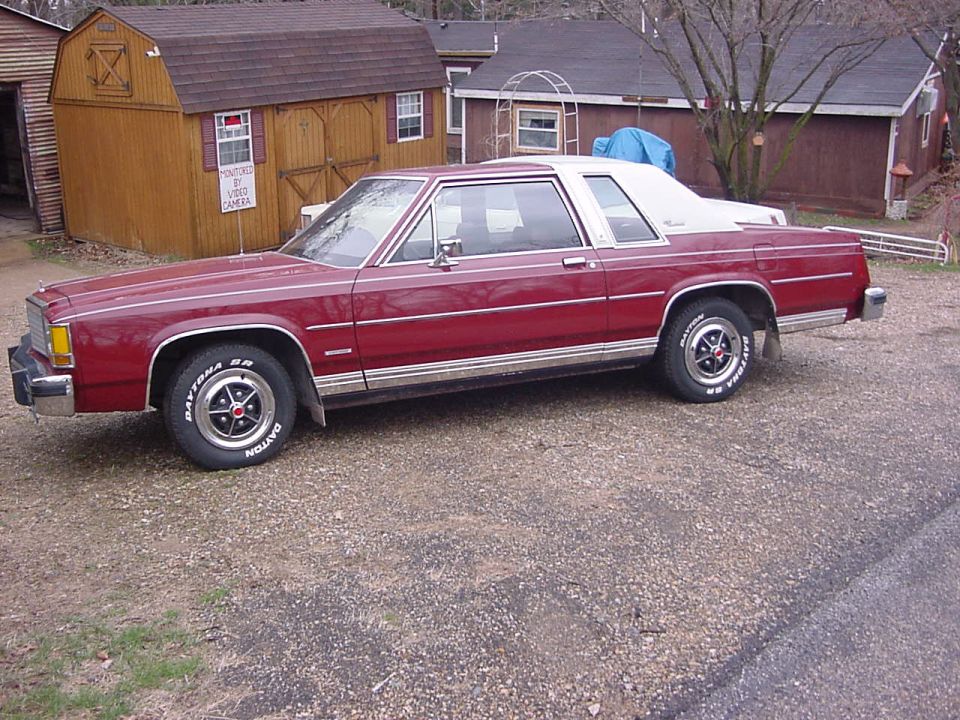 Dark red two-door Ford LTD with white vinyl top and white-letter tires, side view on gravel driveway