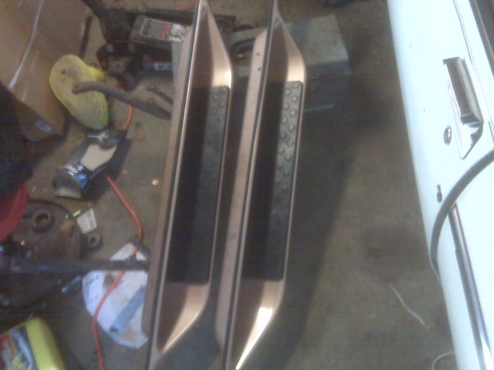 Two metal running boards with black grip pads, one slightly behind the other, with a white vehicle door visible on the right