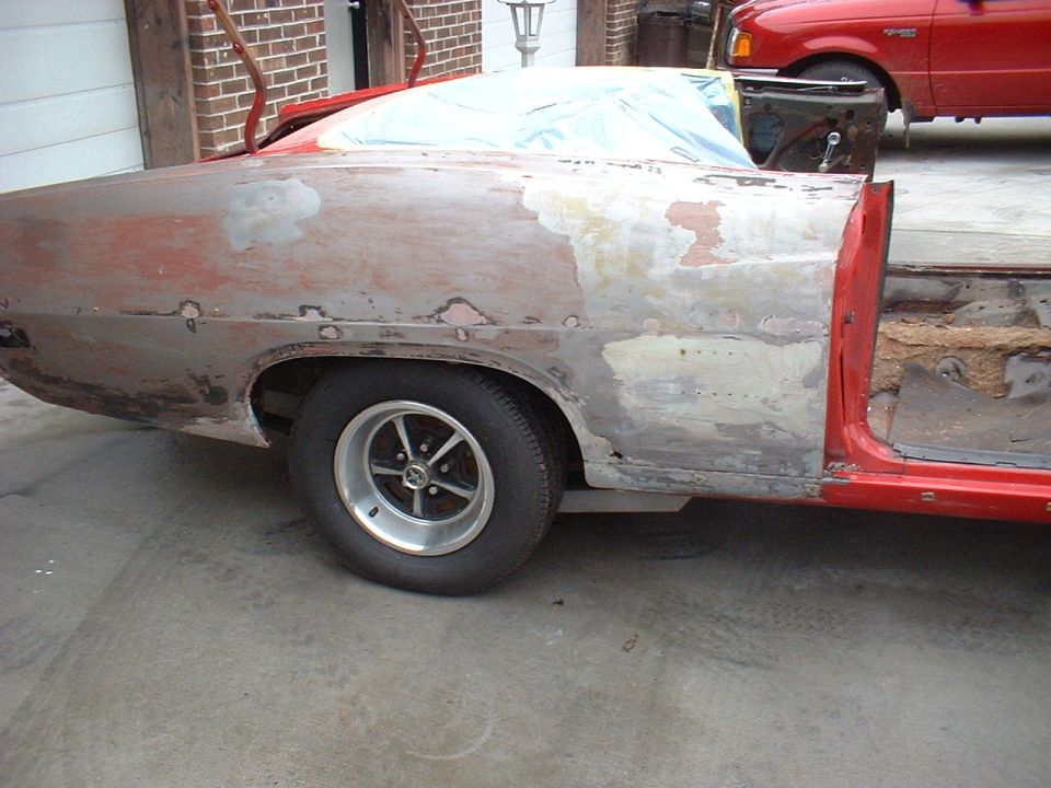 Red 1970 Ford Torino undergoing repairs, showing primer and body filler on the rear quarter panel and door opening