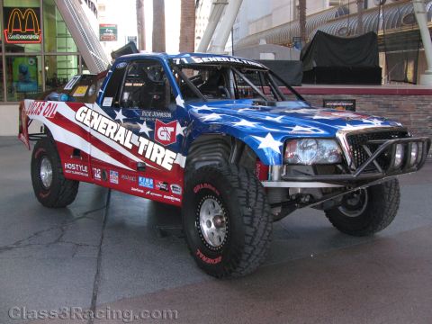 1990 Ford Bronco - red-white-and-blue General Tire off-road race truck numbered 71 is parked on display under a covered walkway by a McDonald's.