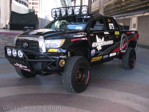 1990 Ford Bronco - black Toyota Tundra off-road race truck numbered 875 with Camburg graphics and a roof light bar sits on display under a canopy.