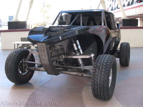 1990 Ford Bronco - black tube-frame off-road race car with exposed front coilover suspension is displayed on a paved plaza under a canopy.