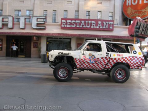 1990 Ford Bronco - White Bronco race truck 317 with red checkered Big Boy graphics drives down a downtown street past a restaurant marquee.