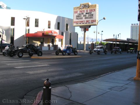 1990 Ford Bronco - line of off-road race buggies drives along a city street past a McDonald's and a Plaza marquee reading Rat Pack Is Back.