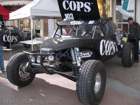 1990 Ford Bronco - black tubular-frame off-road race buggy marked number 103 and COPS, with big coilover shocks and knobby tires, displayed under tents.