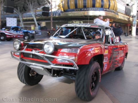 1990 Ford Bronco - red and black off-road trophy truck with a roll cage drives down a downtown street lined with casinos and spectators.