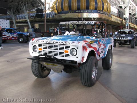 1990 Ford Bronco - white vintage Ford Bronco off-road racer numbered 348 with stars-and-stripes Team B.C. Broncos livery rolls under a covered street.