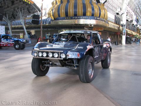 1990 Ford Bronco - dark blue off-road trophy truck with a five-lamp roof light bar and roll cage drives down a covered pedestrian street.