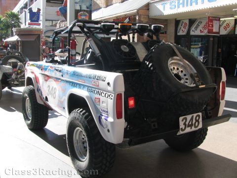 1990 Ford Bronco - Rear three-quarter view of a white off-road Bronco-style race truck numbered 348 with a rear-mounted spare tire and roof light bar.