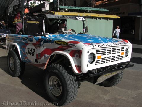 1990 Ford Bronco - white early Ford Bronco off-road racer with American-flag bodywork numbered 348, Team B.C. Broncos, parked on a sunny street.