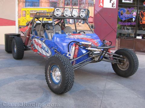 1990 Ford Bronco - blue tubular-frame off-road race buggy with coilover shocks and a roof light bar parked on pavement in front of shops.