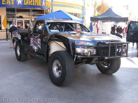 1990 Ford Bronco - black off-road trophy truck with a heavy tube front bumper and roll cage parked on pavement near a blue canopy and storefront.