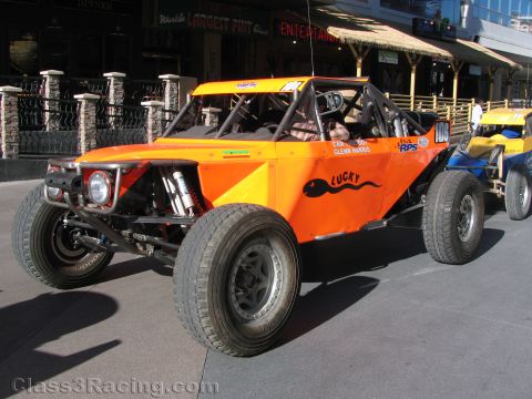 1990 Ford Bronco - orange tubular-frame off-road race buggy labeled Lucky and number 100 parked on a downtown street beside a yellow buggy.