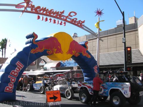 1990 Ford Bronco - blue Red Bull inflatable arch stands under the Fremont East District sign with off-road race vehicles staged on the street.