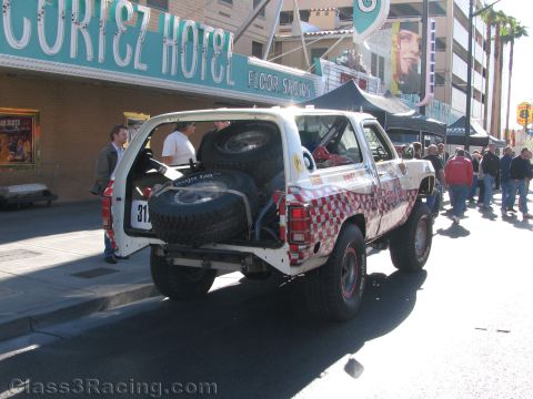 1990 Ford Bronco - Rear view of a white off-road SUV racer with checkered graphics and a rear-mounted spare, parked beside the El Cortez Hotel.