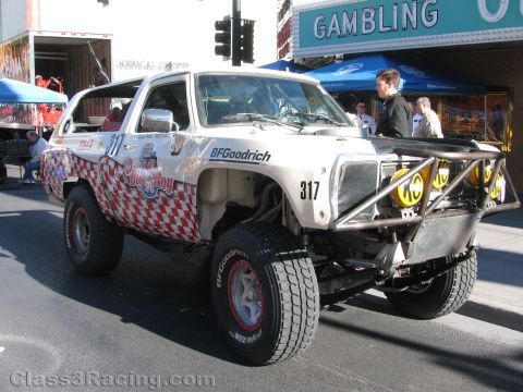 1990 Ford Bronco - white off-road SUV racer with red checkered trim, number 317, and yellow auxiliary lights parked on a street near a Gambling sign.