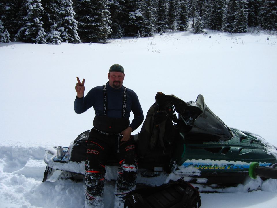 Green snowmobile with 'SUMMIT' visible on side, parked in snow with a man giving peace sign