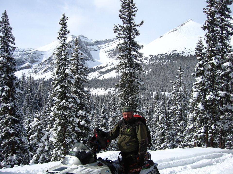 Man standing next to a snowmobile with snow-covered trees and mountains in the background