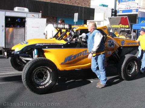 1990 Ford Bronco - yellow tubular-frame off-road race buggy numbered 154 sits in a pit area with Goodyear and Lucas Oil banners; a man stands beside it.