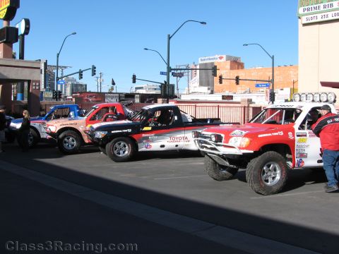 1990 Ford Bronco - Several off-road trophy trucks, including red Toyota-branded ones, staged at a sunny downtown intersection near a Bank of America.