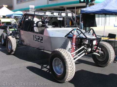 1990 Ford Bronco - bare aluminum-bodied tube-frame off-road buggy numbered 1522 with red coilover shocks parked on a downtown street.