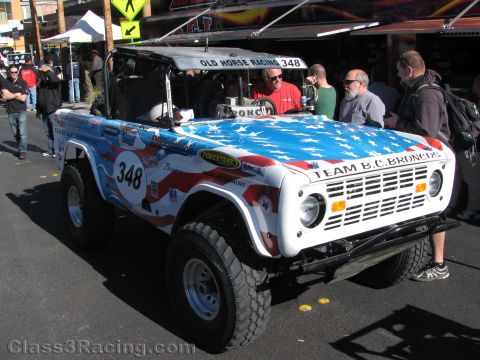 1990 Ford Bronco - white American-flag Ford Bronco off-road racer numbered 348, Team B.C. Broncos, parked on a street as spectators gather around.