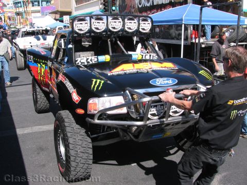1990 Ford Bronco - black Ford off-road race truck with Monster Energy and AEM graphics and a five-lamp roof light bar being pushed along a street.