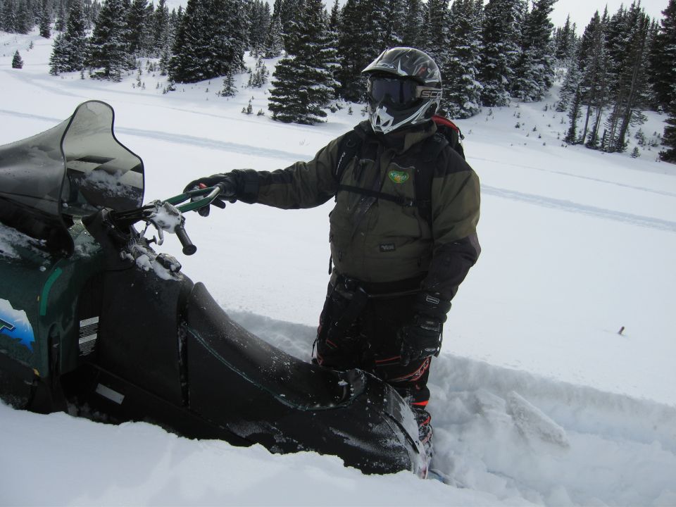 Green snowmobile partially buried in snow with a person in a helmet and goggles standing next to it