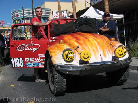 1990 Ford Bronco - flame-painted off-road VW Beetle race car numbered 1188 with KC lights and roll cage, displayed curbside with two men.
