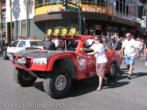 1990 Ford Bronco - red trophy-style off-road race truck numbered 6084 with roof light bar rolls down a street as pedestrians walk alongside.