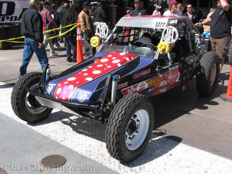 1990 Ford Bronco - patriotic-themed open desert race buggy numbered 1377 with smiley-face driving lights, surrounded by spectators.