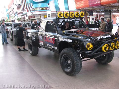 1990 Ford Bronco - black trophy-style race truck with a roof rack of KC lights and Yokohama branding parked under a covered Las Vegas street.