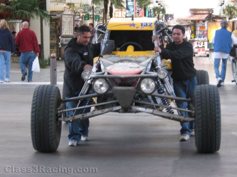 1990 Ford Bronco - Two men push an open tube-frame desert race buggy numbered 132 down a pedestrian street lined with shops.