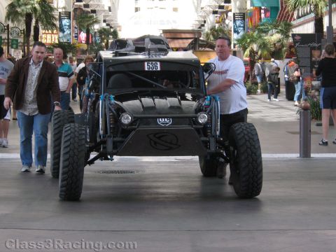 1990 Ford Bronco - man pushes a lifted off-road race car numbered 536 with oversized tires along an indoor covered walkway.