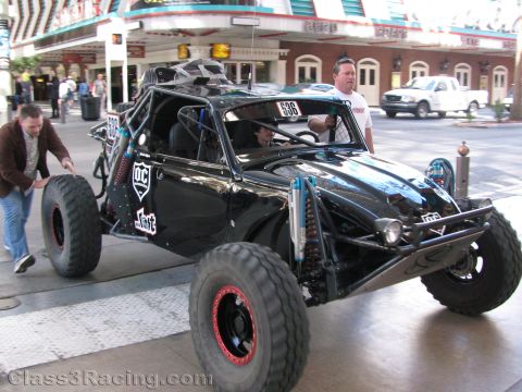 1990 Ford Bronco - black open tube-frame desert race buggy with red beadlock wheels is rolled along a street as people walk by.