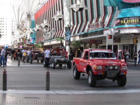 1990 Ford Bronco - red off-road race truck leads a group of race vehicles along the Fremont Street Experience as spectators look on.