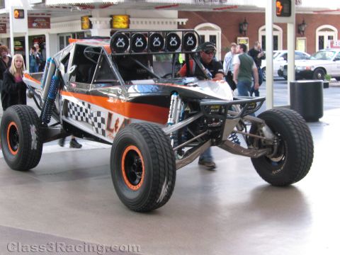 1990 Ford Bronco - black-and-orange open desert race buggy with checkered graphics and a roof light bar parked on a covered walkway.