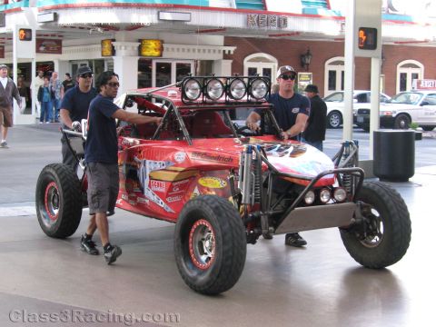 1990 Ford Bronco - Crew members push a red open desert race buggy with BFGoodrich graphics and roof lights along a covered Las Vegas street.