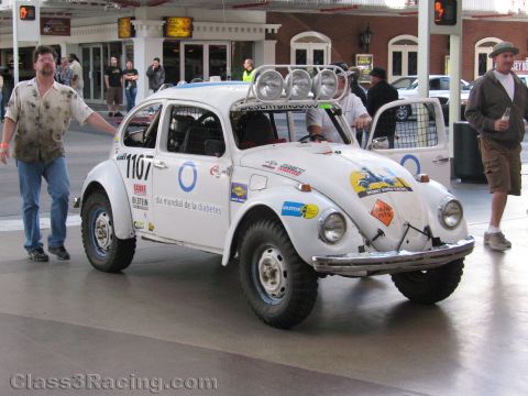 1990 Ford Bronco - white off-road race VW Beetle numbered 1107 with roof driving lights is rolled along a street between two men.