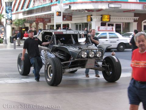 1990 Ford Bronco - black open tube-frame desert race buggy numbered 124 with BFGoodrich branding rolls past spectators on a city street.