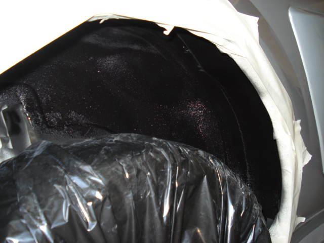 Wheel well interior covered in black undercoating and plastic wrap. White tape masks the surrounding body panel