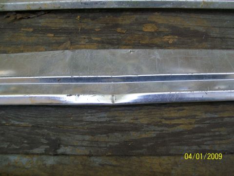 1972 Ford Ranchero - A close-up view of a dented and scratched chrome trim piece, likely a vehicle part, on a wooden surface.