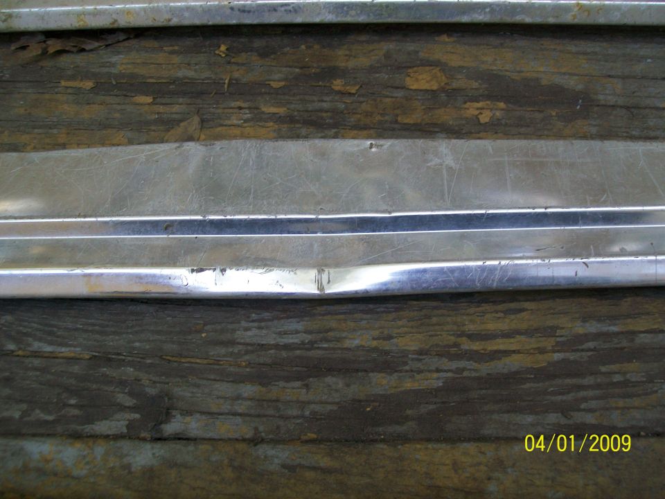 Damaged chrome trim piece with visible dent and scratches, resting on weathered wood